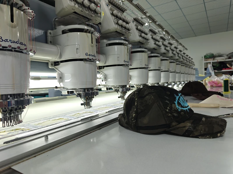 Dark camouflage cap with turquoise embroidery on the table beside multihead embroidery machines